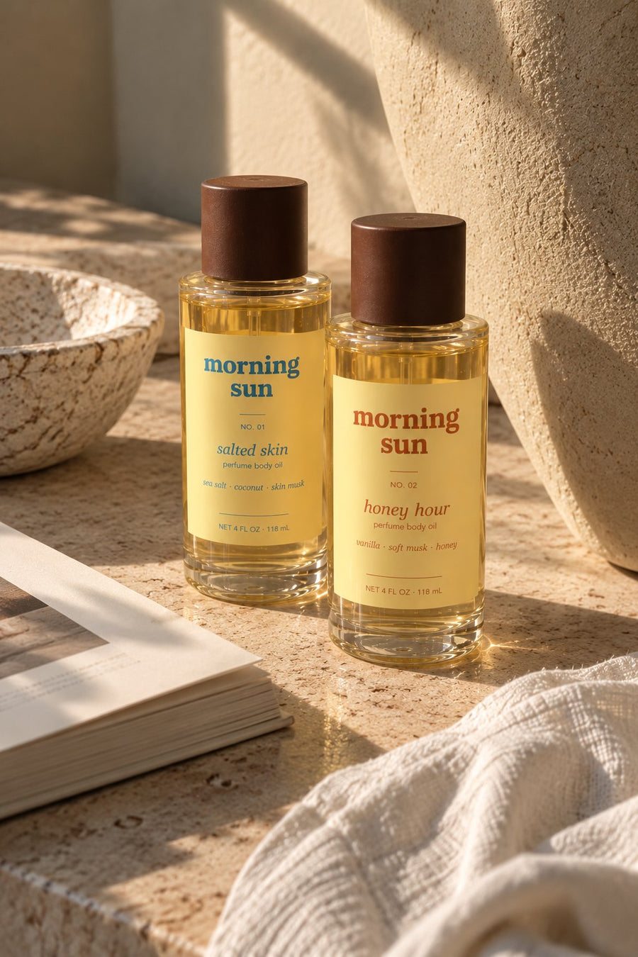 Salted Skin and Honey Hour perfume body oils on a stone vanity in warm morning light, with linen and a ceramic bowl