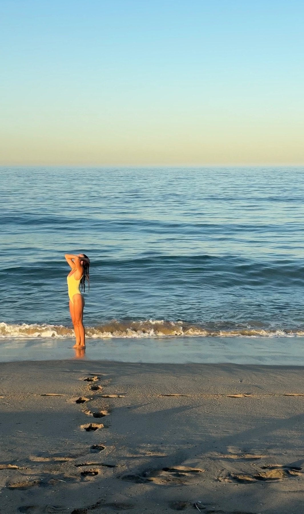 Paige in a yellow swimsuit at the water’s edge at sunrise, wringing out her wet hair
